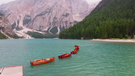 Drone orbit slow across Braies lake in Dolomites with line of red boats over clear water