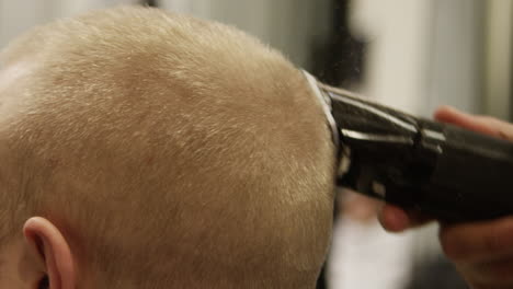 Male-barber-giving-a-blonde-caucasian-man-a-haircut-at-the-Barbershop,-by-shaving-his-head-with-an-electronic-razor-in-slow-motion