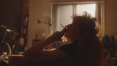 joven pensativo sentado en un sillón, fumando un cigarrillo en una habitación desordenada