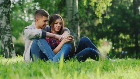 Mann-Und-Frau,-Die-Sich-Umarmen,-Sitzen-Auf-Dem-Gras-Im-Park-Und-Schauen-Auf-Den-Handybildschirm,-Kommunizieren-Sm