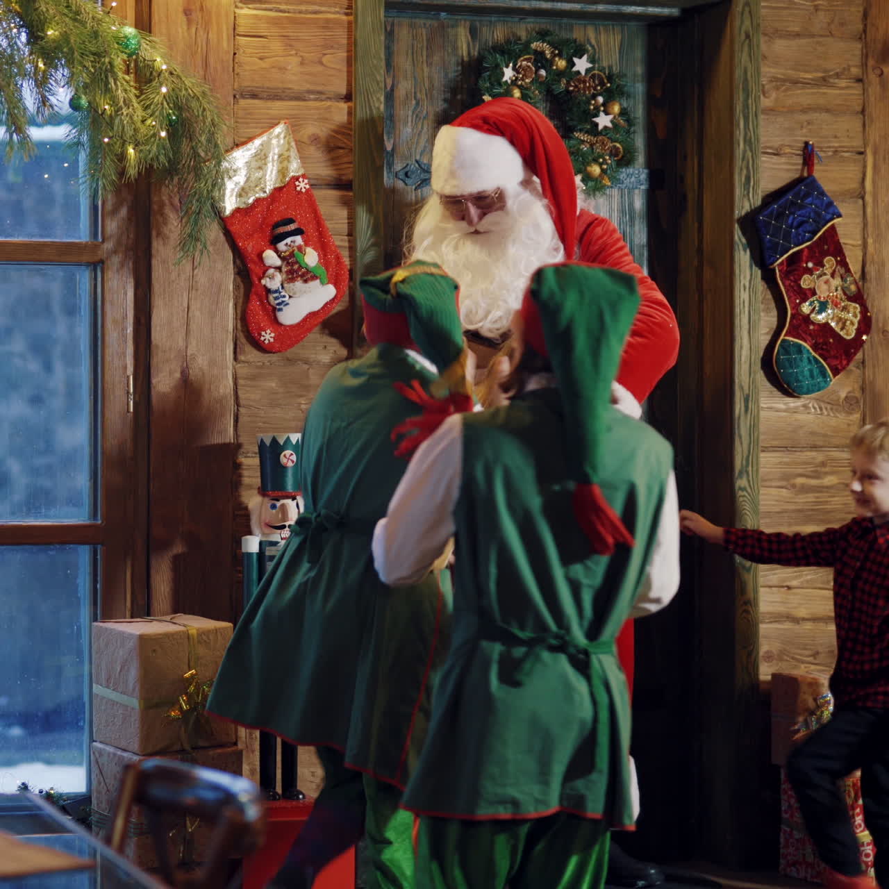 Santa Claus opens the door and comes inside his residence. Happy children and fairy elves running to Santa in Christmas decorated room.