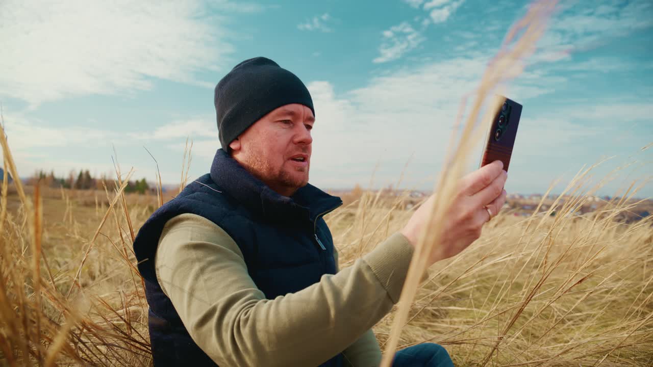 A person capturing a moment on a smartphone amidst a field of tall grass under a blue sky, showcasing a sense of connection with nature while engrossed in their device, reflecting modern life's blend of technology and outdoor experience