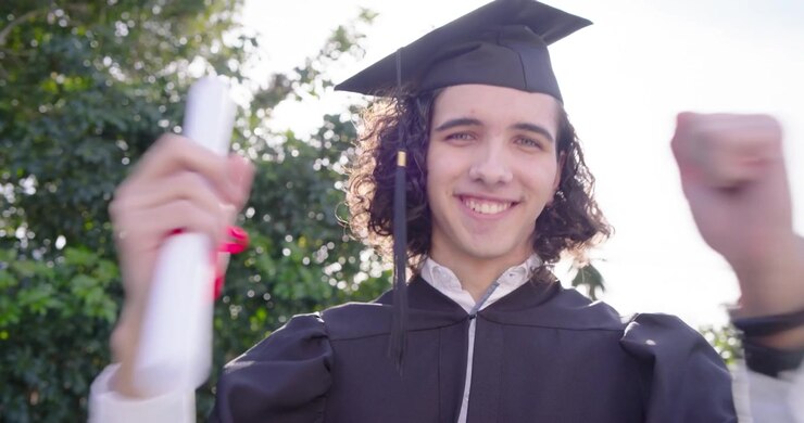 Laureato con tocco e toga che alza e ruota un diploma con nastro rosso, alzando il pugno in segno di gioia, in giardino