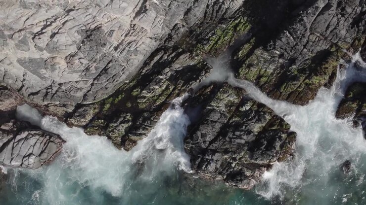 Drammatica prospettiva dall'alto di potenti onde marine che si infrangono su rocce scure e affilate, evidenziando l'energia grezza e l'erosione della costa. 4K Dlog.