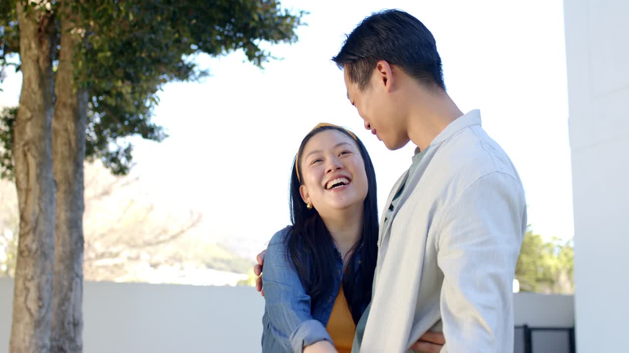 多様なカップルが、テラスの低い白い壁のそばで、通知後にスマートフォンを共有し、笑い合い、抱き合っています。