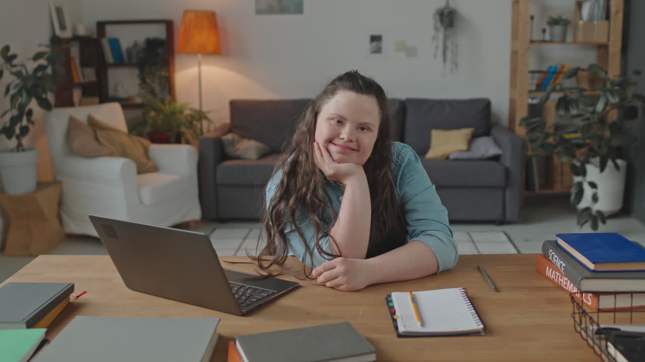 A young woman with Down syndrome is sitting at a table with a laptop and books