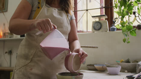 mid section of female caucasian potter pouring paint from a jug on pot at pottery studio