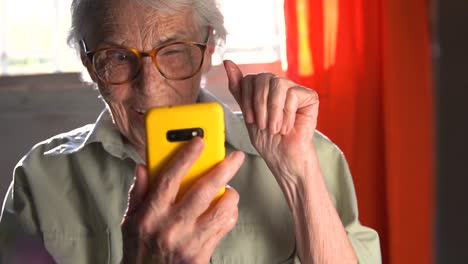 un primer plano de una feliz anciana con gafas, involucrada en una llamada de video en su teléfono inteligente con un caso amarillo brillante