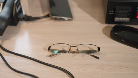 a close-up view of a hand wearing a black glove dropped a pair of glasses on a wooden desk, with phone on the table and other equipment