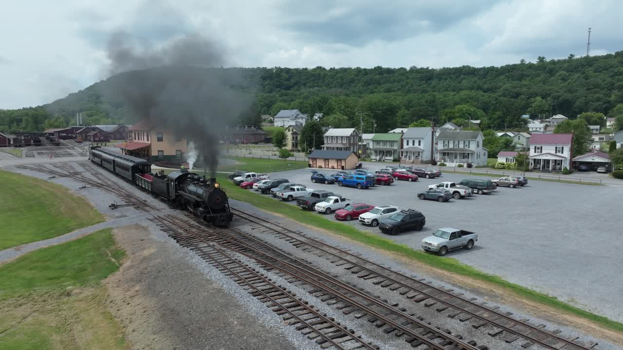 A narrow gauge steam train puffs clouds of smoke as it moves past a small town. The engine is surrounded by parked cars and charming houses under a bright sky