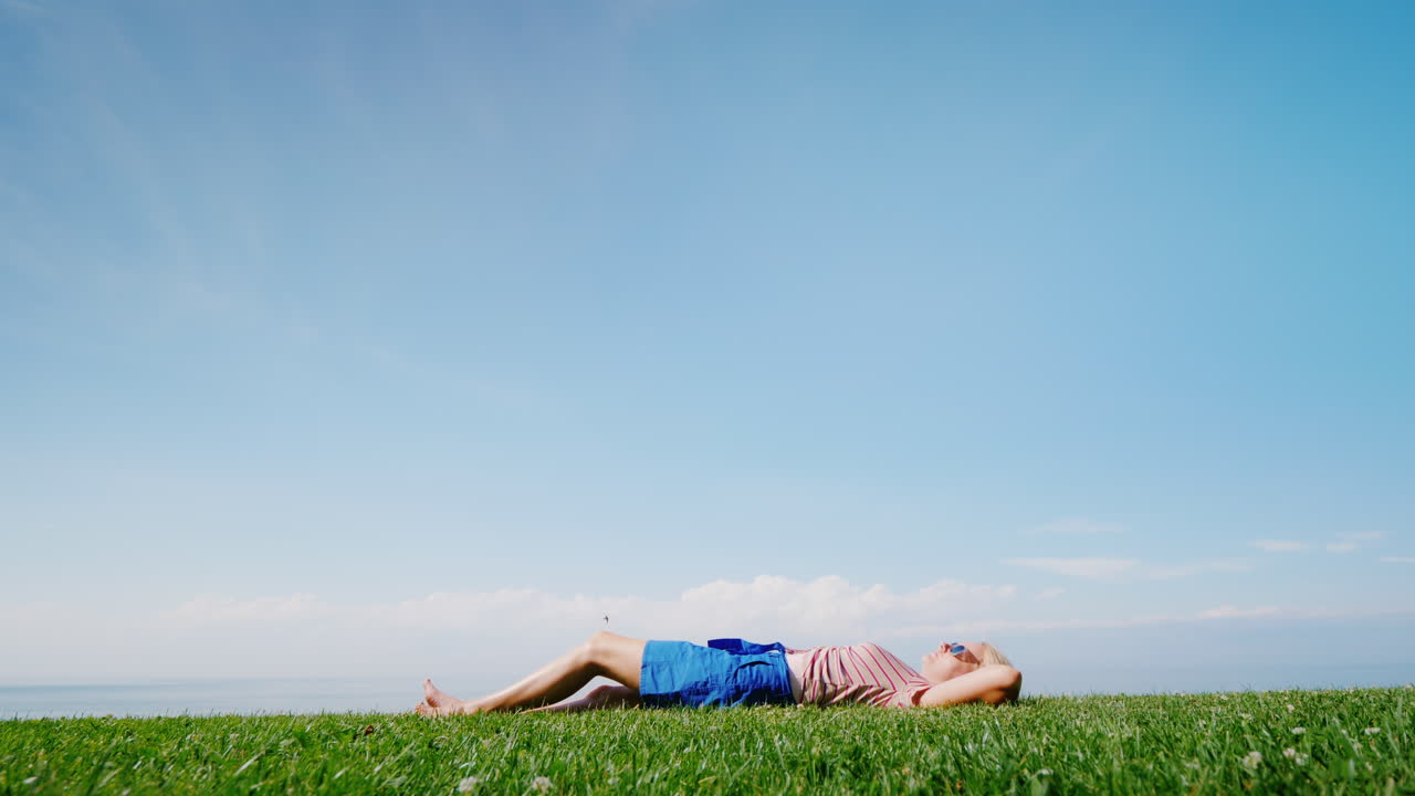 한 젊은 여성이 푸른 하늘을 배경으로 푸른 잔디에 누워 따뜻함과 여름을 즐긴다