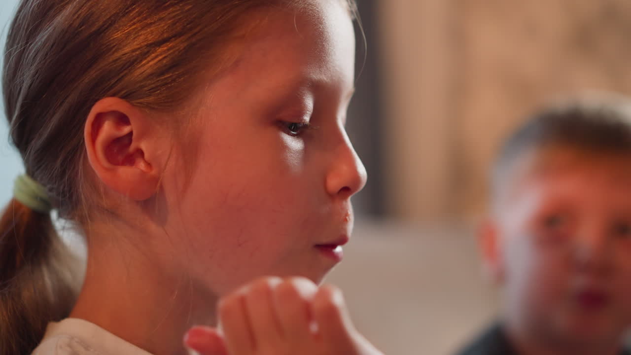 closeup of girl child seated indoors eating fried chips dipped into sauce playfully with ketchup stain near lips while slightly blurred view of young boy in background