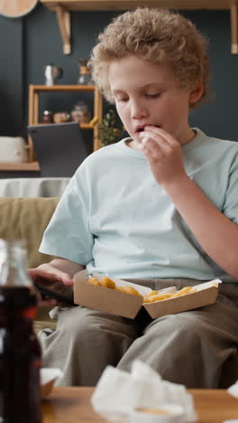 young boy eats fast food while using smartphone at home