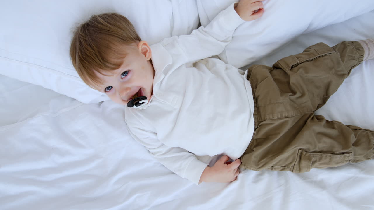 Beautiful smiling Caucasian baby with pacifier in mouth. Kid lying on the bed smiles adorably. Top view.
