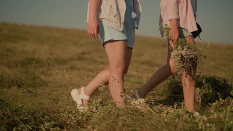 close up of two women walking through grassy field, one holding bouquet of wildflowers, sunlight highlights their legs and field, capturing natural beauty and carefree vibe of outdoor activity