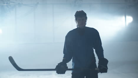 Toma-De-Retrato-De-Un-Jugador-De-Hockey-Masculino-Concentrado-Parado-En-La-Arena-De-Hielo,-Sosteniendo-Un-Palo-Y-Mirando-La-Cámara