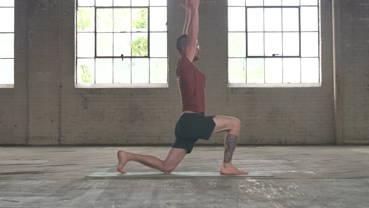 Man in an industrial warehouse practicing yoga, the pose of low lunge with side stretches.