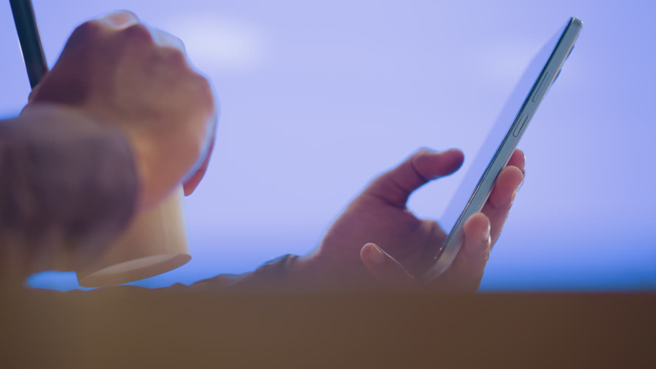 Person holding smartphone in one hand with blank screen visible while sipping drink with other hand, warm indoor atmosphere enhanced by soft bokeh lighting, conveying relaxed digital lifestyle moment