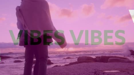 animation of good vibes text over couple wrapped in blanket standing on beach with pink tint