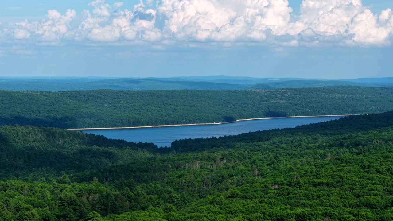 Drone footage reveals Quabbin Reservoir's receding blue waters and exposed shoreline, surrounded by dense green forest under dramatic clouds, highlighting drought impact in daylight.