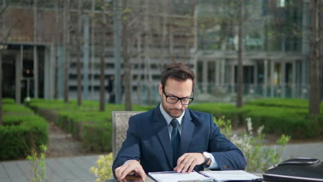 caucasian businessman man in suit doing paperwork in a park, watching phone while sitting by the table in city park by the office - orbit shot