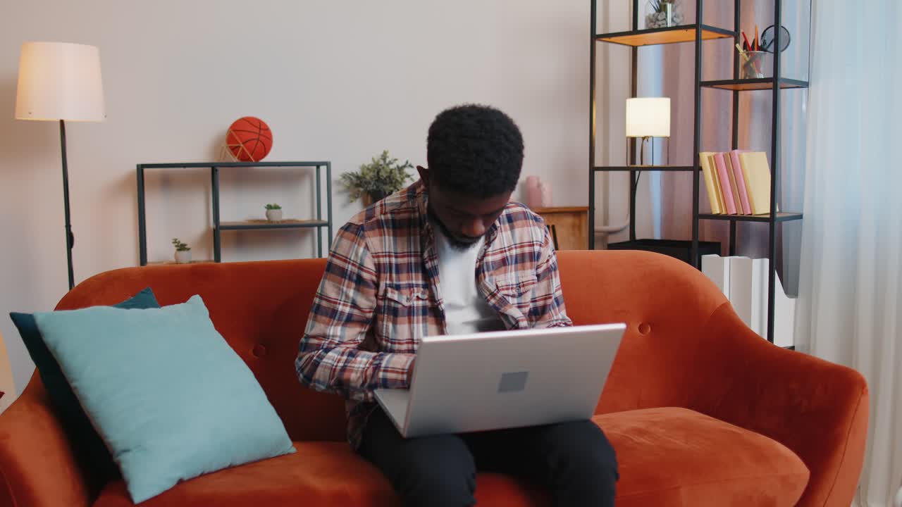 African american man sitting on sofa opening laptop pc starting work online in living room at home
