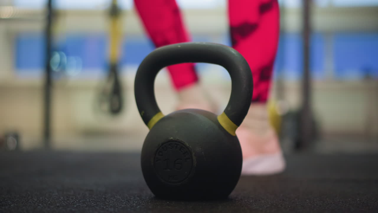 Kettlebell sul pavimento della palestra vicino all'atleta che si avvicina all'impugnatura preparandosi per lo swing, leggings rosa acceso e scarpe da ginnastica, pavimento in gomma, attrezzatura sfocata sullo sfondo, illuminazione interna calda, allenamento concentrato.