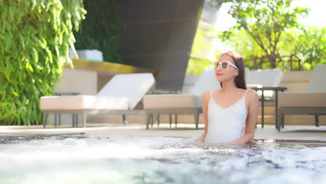 Schicke-Asiatische-Frau-In-Sonnenbrille-Und-Badeanzug,-Die-An-Sonnigen-Sommertagen-Im-Jacuzzi-Eines-Schwimmbades-Genießt,-Zeitlupe