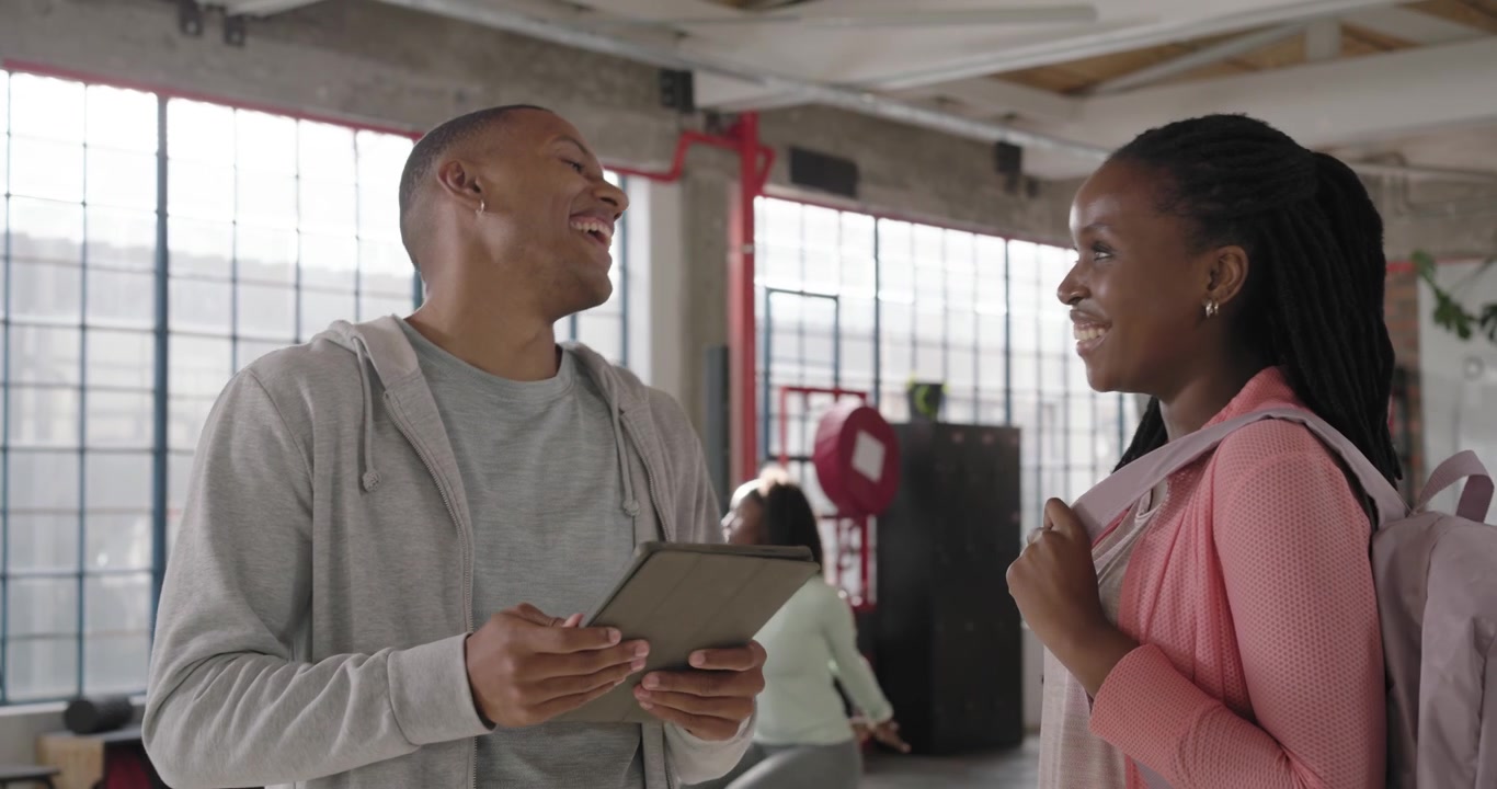Coach scanning tablet and showing plan, African American woman listening with pink backpack at gym
