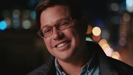 portrait young caucasian man on rooftop at night wearing glasses smiling happy enjoying urban nightlife with bokeh city lights in background