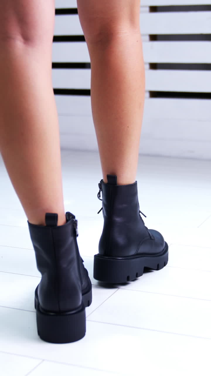 Female Caucasian model wearing mini skirt walks in the room demonstrating black boots on tractor soles with laces. White backdrop. Vertical video