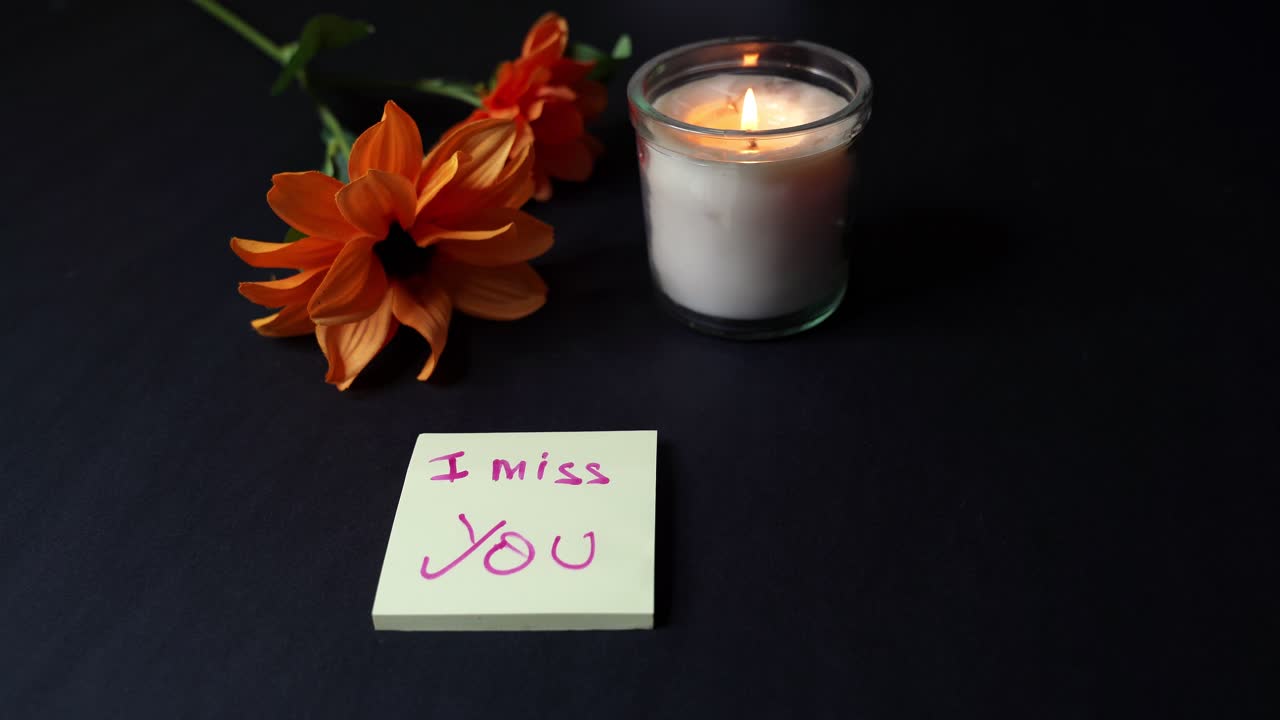 Oversigtsbillede af hænder, der skriver “Jeg savner dig” på en sticky note med en pink tusch, varmt stearinlys og orange blomster på et mørkt bord, romantisk beskedkoncept med kopiplads, natstemning