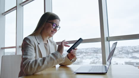 happy smiling young woman wearing glasses dial telephone number. long-haired lady sits at desk with laptop near the big window.