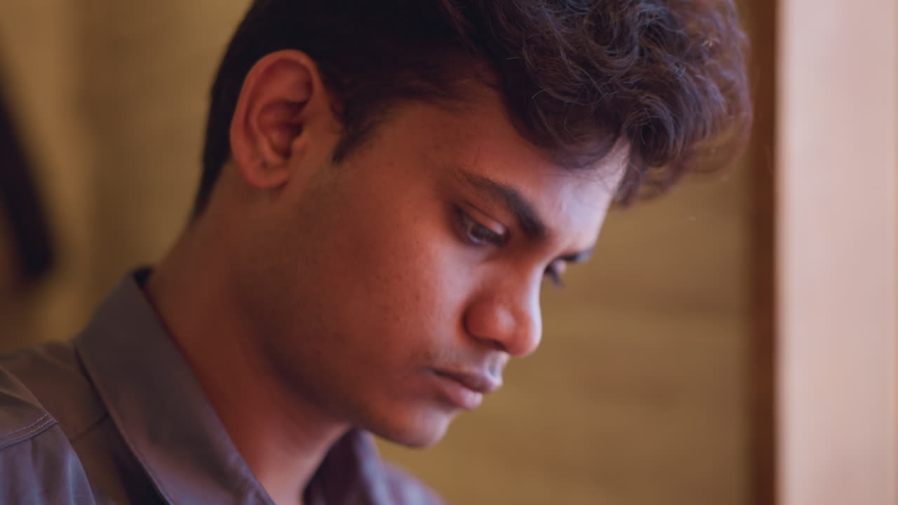 Close-up side profile of young man in natural light looking out window with serious expression, reflecting deep thought or contemplation, captured in quiet indoor setting with soft brown background tones