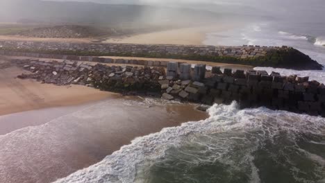 Vista-Aérea-De-Las-Olas-Rompiendo-Contra-La-Pared-Rocosa-En-La-Playa-De-Arena-Con-La-Desembocadura-Del-Río-En-Nazaré,-Portugal---Rio-Alcoa,-Leira