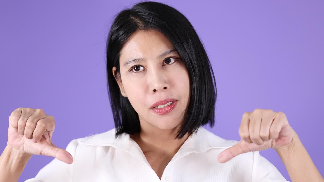 Serious woman in white shirt shows thumbs down, expressing disapproval, against a purple background