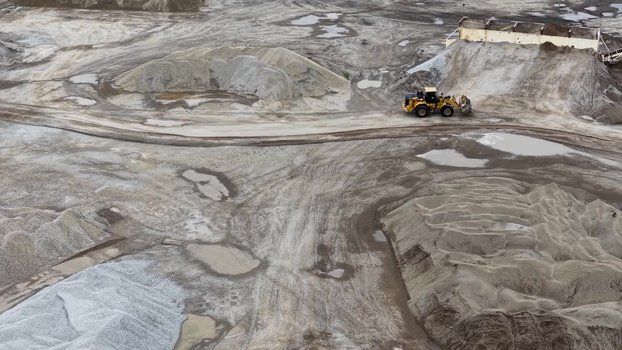 Low aerial pass over pale gravel mounds and winding tire tracks across the quarry floor