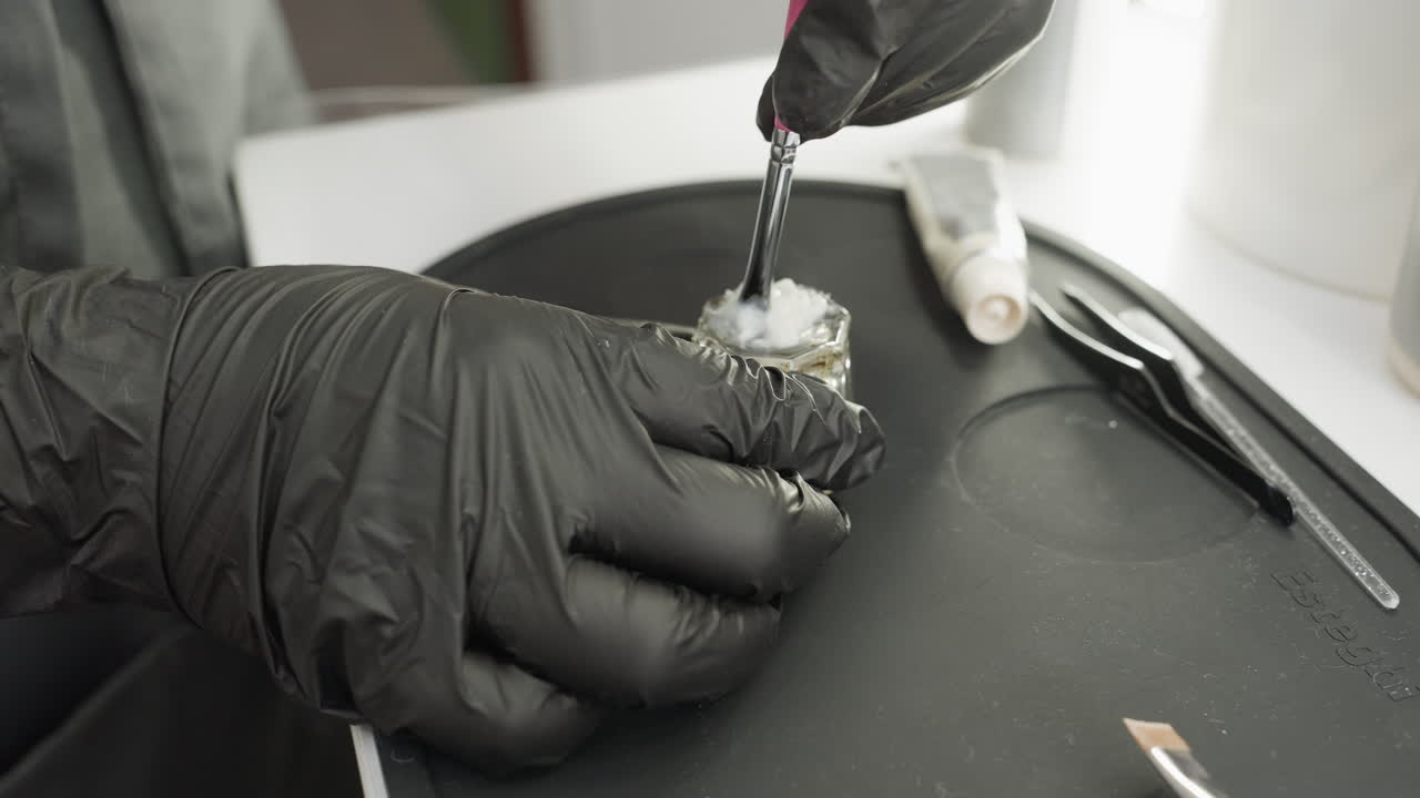 Close-up of technician wearing black gloves using brush to mix thick white cream inside small transparent glass jar placed on black silicone tray with beauty tools arranged around work area