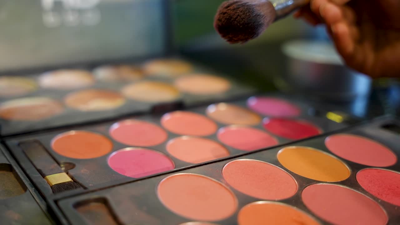 Pennello da trucco tenuto sopra una palette cosmetica colorata con diverse tonalità di blush e cipria disposte in un vassoio.