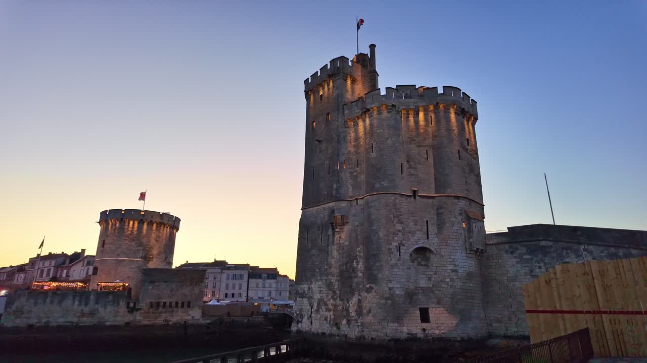Camera moves forward toward Tour de la Chaîne in La Rochelle port, towers lit against sunset sky - France