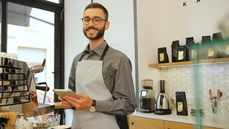 barista caucásico con barba y anteojos usando la tableta, luego mira la cámara y sonríe en una cafetería