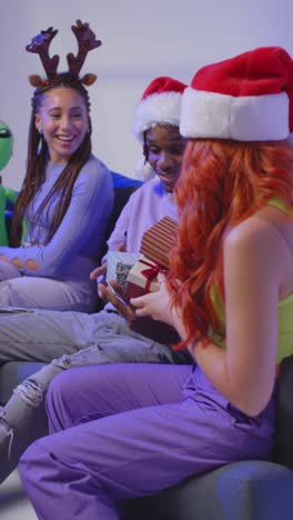 vertical video studio shot of gen z friends giving and opening presents for christmas sitting on sofa wearing santa hat and reindeer antlers