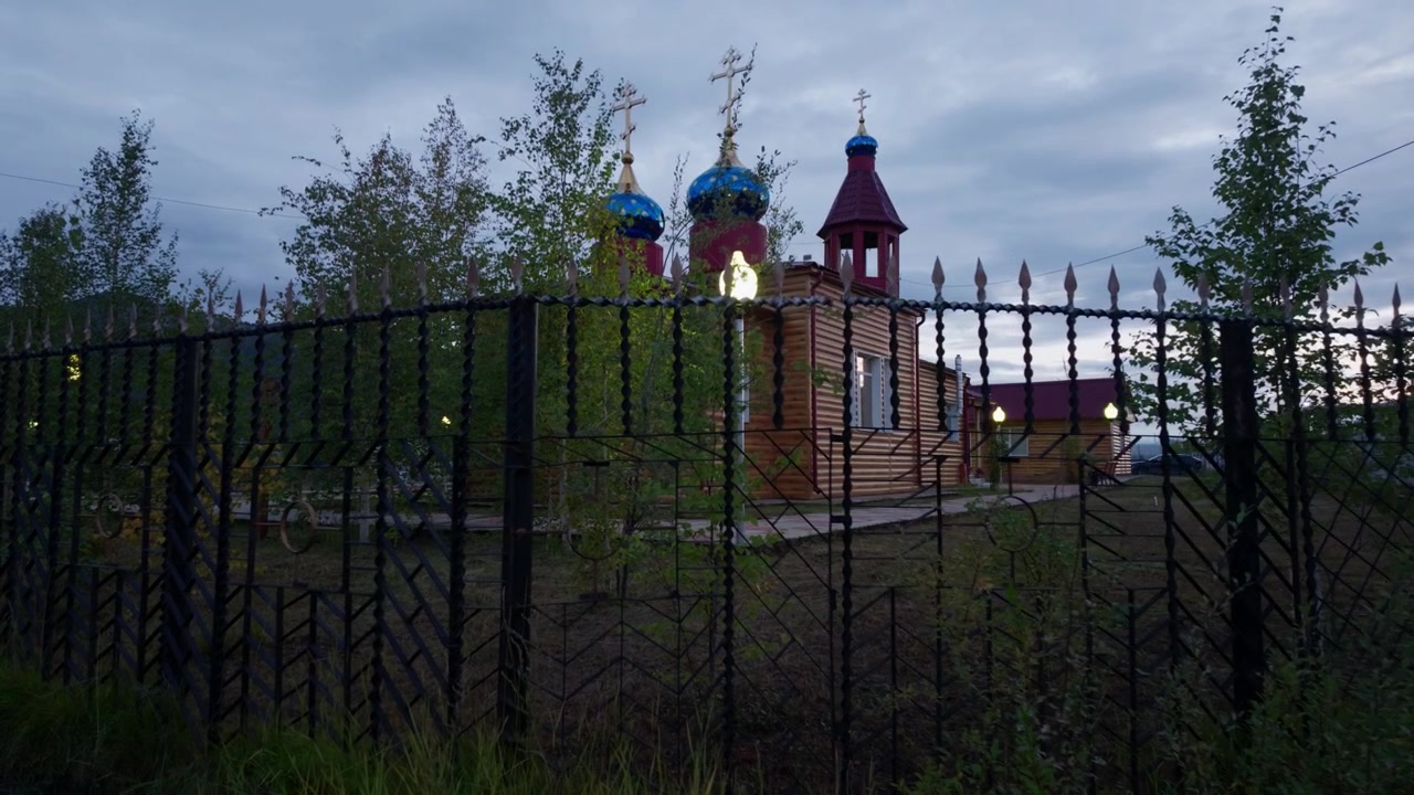 A wooden Orthodox church with blue and red domes and golden crosses stands behind a black metal fence, surrounded by trees under a cloudy twilight sky as the camera slowly pans right.