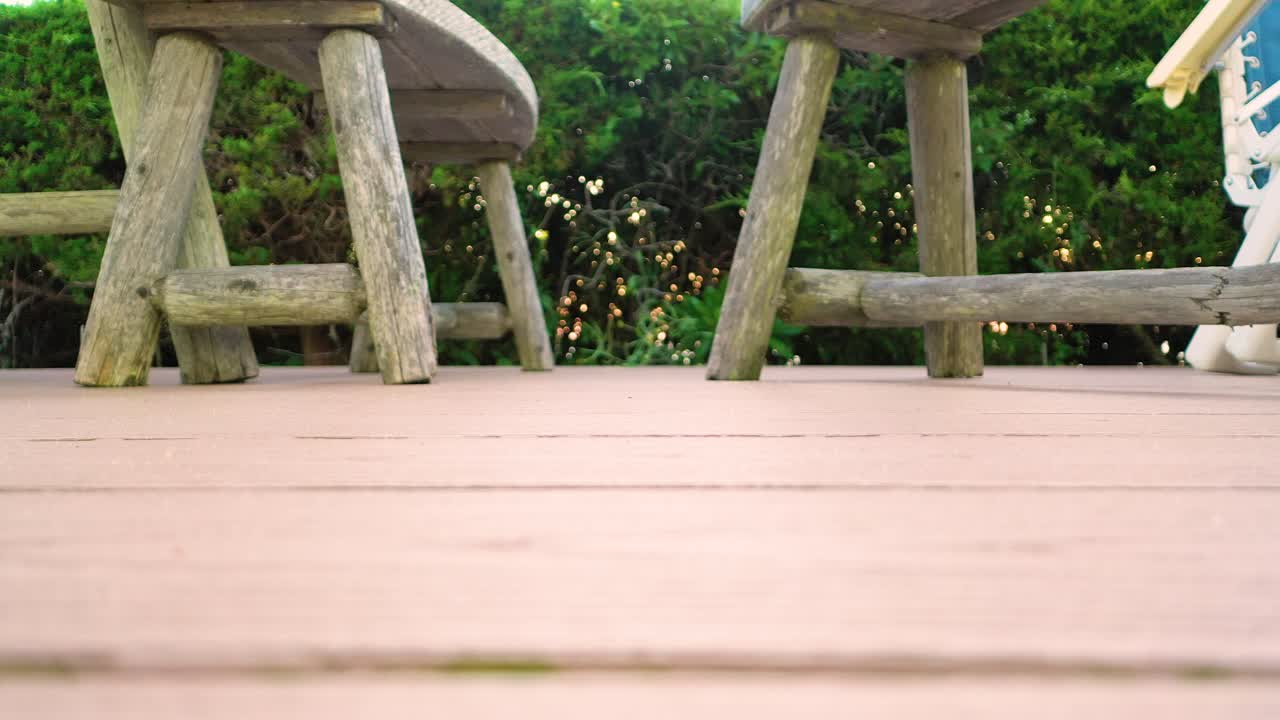 Slow pan across a wooden deck with rustic outdoor chairs and soft greenery in the background. Shallow depth of field and warm light create a calm, cozy atmosphere, ideal lifestyle or backyard b-roll