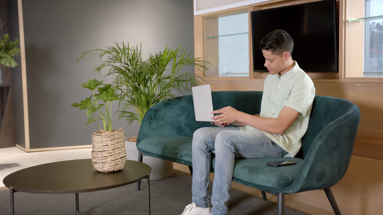 Sitting on couch, man using laptop and smartphone in modern office setting, copy space