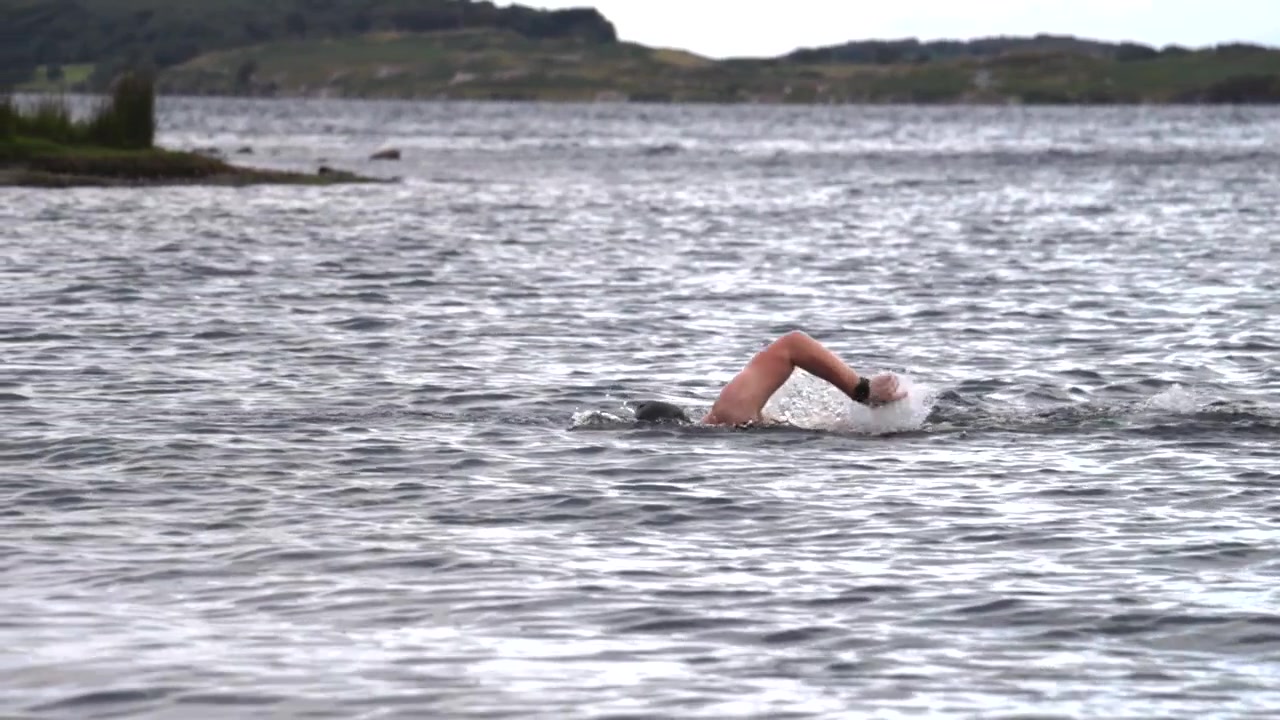 Open water swimmer in the UK filmed from the side in slow motion, freestyle stroke with splashes and ripples on a lake or sea during outdoor endurance training.