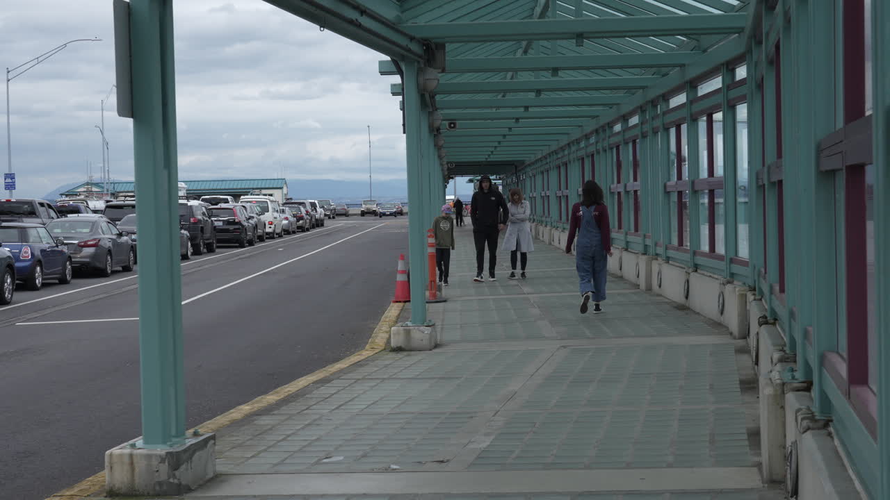 People walking towards the Clinton Ferry Terminal along a covered walkway, while vehicles wait in line to board the ferry on an overcast day.