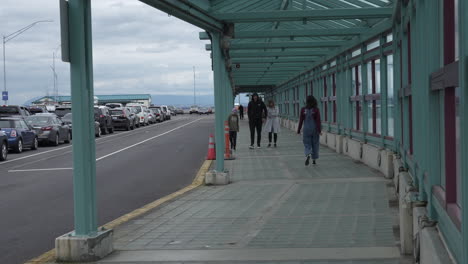 people walking towards the clinton ferry terminal along a covered walkway, while vehicles wait in line to board the ferry on an overcast day.