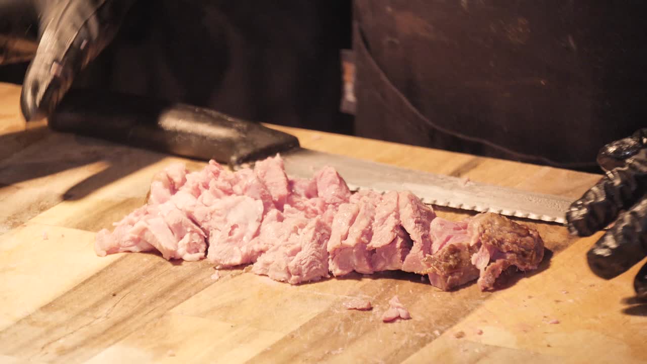 Uno chef con guanti neri taglia pezzi di prosciutto e carne con una mannaia e un coltello su un tagliere di legno.