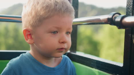 Little-boy-sits-in-poma-lift-cabin-riding-against-mountains